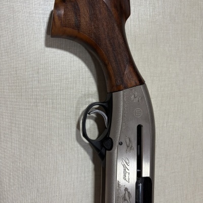 Espingarda de caça com empunhadura de madeira castanha e corpo metálico prateado com gravações e texto.