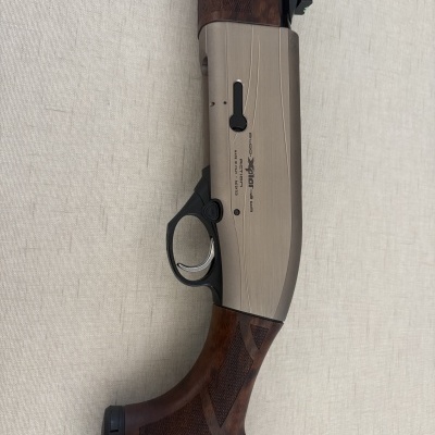 Espingarda com madeira castanha texturizada e corpo metálico cinzento com texto em fundo branco.