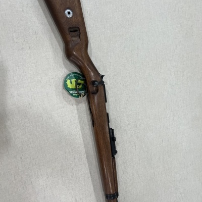 Espingarda com coronha de madeira castanha e detalhes metálicos pretos sobre fundo bege