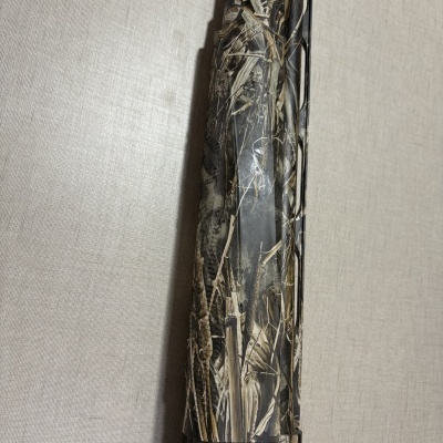 Arma de fogo camuflada com padrão de folhas secas e ramos sobre fundo bege texturizado.