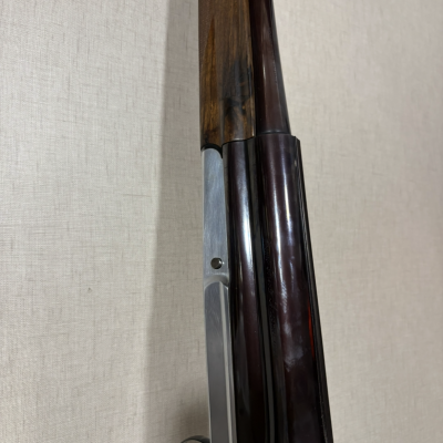 Espingarda de canos paralelos com madeira castanha e metal escuro apoiada numa parede bege.