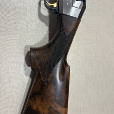 Espingarda com coronha de madeira escura, gatilho dourado e metal prateado encostada numa parede bege