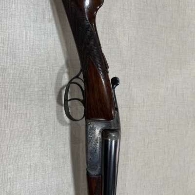 Espingarda de canos paralelos com cabo em madeira castanha e metal escurecido gravado sobre fundo cinzento