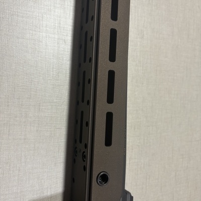 Objeto metálico cinza escuro com perfurações e parafusos sobre fundo tecido cinza