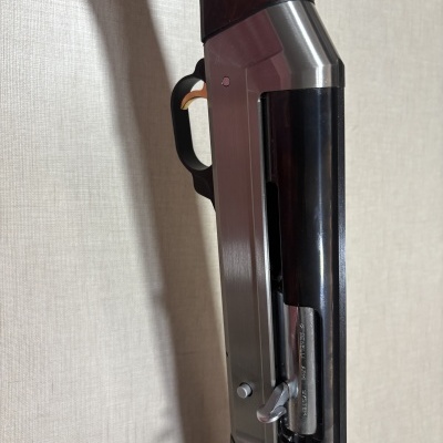 Espingarda de caça com detalhes em madeira, metal prateado e preto, gatilho dourado, contra fundo neutro.