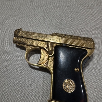 Pistola dourada com cabo preto e gravações decorativas sobre superfície cinzenta.
