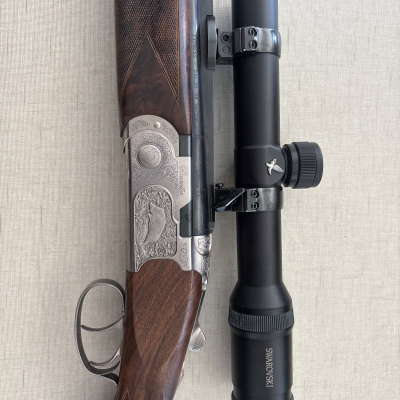 Rifle de madeira com metal gravado e luneta óptica preta SWAROVSKI sobre tecido bege.