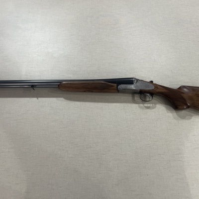 Espingarda de canos duplos com coronha em madeira castanha sobre fundo cinza.