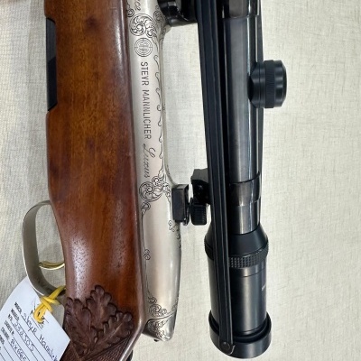 Rifle com coronha em madeira trabalhada e acabamento metálico prateado com gravações e mira telescópica preta.