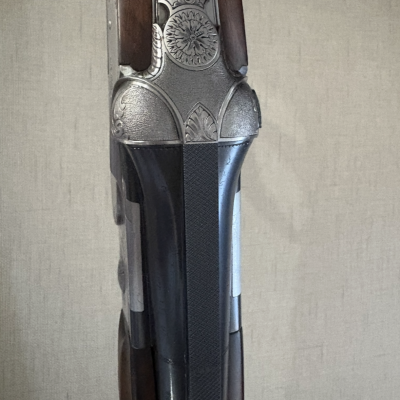 Parte superior de espingarda com metal gravado, madeira escura e correia preta contra fundo bege