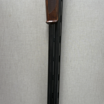 Espingarda com cano preto e punho em madeira castanha, contra parede cinza