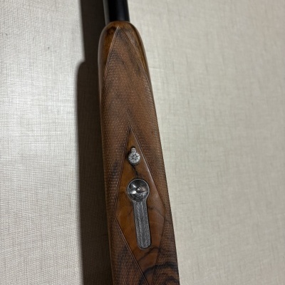 Empunhadura de arma de fogo em madeira trabalhada com detalhes metálicos contra uma parede texturizada bege.