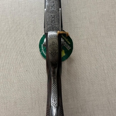 Espingarda com coronha em madeira escura e metal gravado, etiqueta verde visível, sobre superfície bege