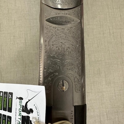 Parte superior metálica de arma com gravações e etiqueta SPORTARCO pendurada em fundo bege