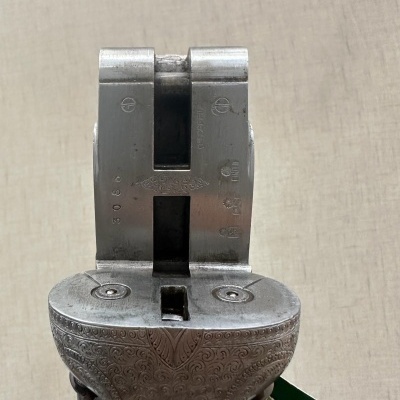 Parte superior de arma de fogo antiga em metal prateado gravado, contra fundo bege.