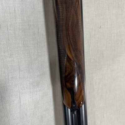 Arma de caça com empunhadura de madeira castanha e canos pretos sobre fundo bege