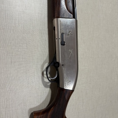 Espingarda com coronha de madeira escura e corpo de metal prateado gravado com aves e texto, sobre fundo bege.