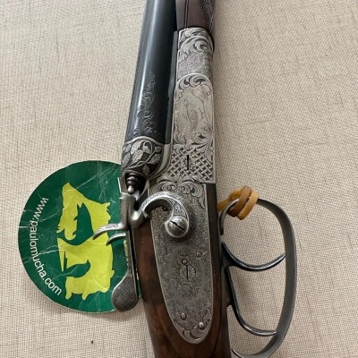 Espingarda dupla com gravações em metal e madeira, etiqueta verde com texto e logo amarelo