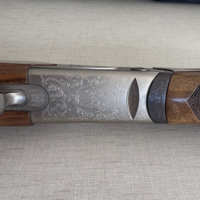 Rifle de caça com coronha de madeira e gravuras no metal, sobre superfície cinza claro.