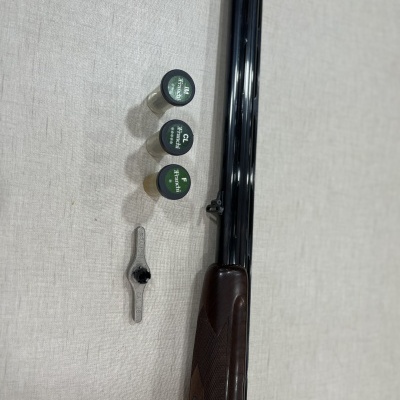 Espingarda de canos duplos com três cartuchos rotulados e uma ferramenta metálica sobre fundo claro