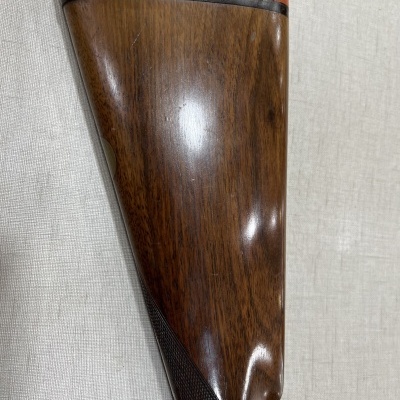 Cabo de espingarda em madeira com borracha vermelha e detalhe preto sobre tecido bege.