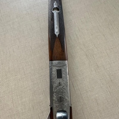 Espingarda de madeira escura com detalhes metálicos gravados, sobre tecido bege.