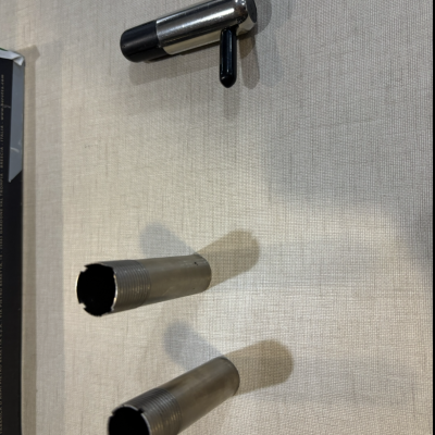 Três acessórios metálicos sobre superfície bege, dois cilíndricos com roscas e um com eixo cromado e cruzado preto.