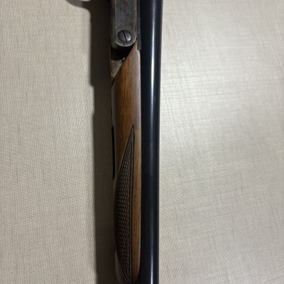 Espingarda de cano duplo em madeira e aço escurecido, vista lateral sobre fundo claro