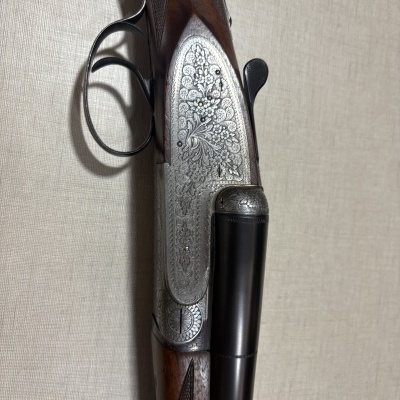 Espingarda dupla de caça com madeira escura e metais gravados em fundo bege.