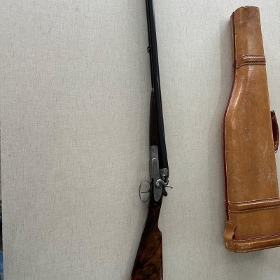 Espingarda de canos duplos com coronha de madeira escura e estojo de couro castanho pendurados na parede