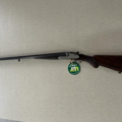 Espingarda de cano duplo com madeira escura e metal preto na parede clara.