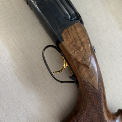 Espingarda com coronha em madeira escura e gatilho dourado sobre tecido bege.