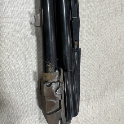 Arma de cano duplo preto com mecanismo metálico cinzento sobre tecido claro.