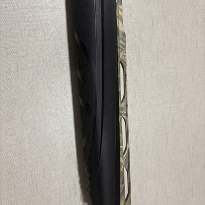 Espingarda em padrão camuflado com guarda-mão preto texturizado encostada numa parede clara