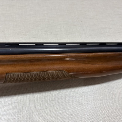 Parte de arma de fogo com guarda-mão em madeira castanha e tubo preto com ranhuras, sobre superfície clara.
