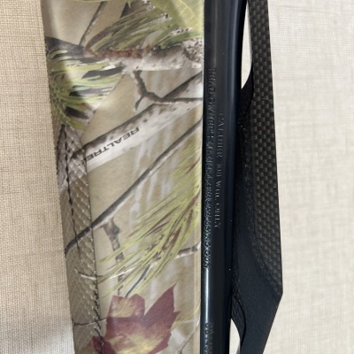 Arma de caça com padrão camuflado e acabamento preto, sobre superfície bege texturizada