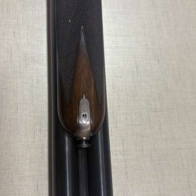 Detalhe inferior de coronha em madeira escura com metal prateado gravado em cano duplo negro