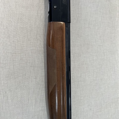 Parte dianteira de espingarda com coronha em madeira castanha e corpo metálico preto sobre fundo cinza