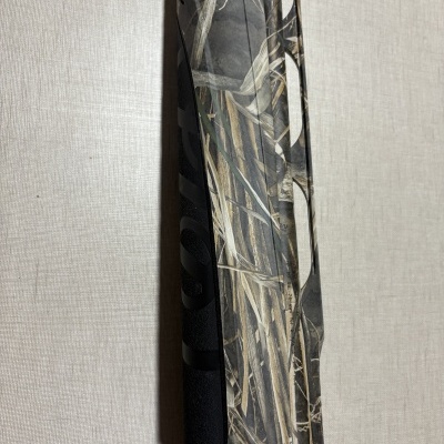 Detalhe de arma de caça camuflada com padrão vegetal e texto parcial.