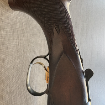 Cabo de espingarda em madeira escura com textura e detalhes em metal preto e dourado.