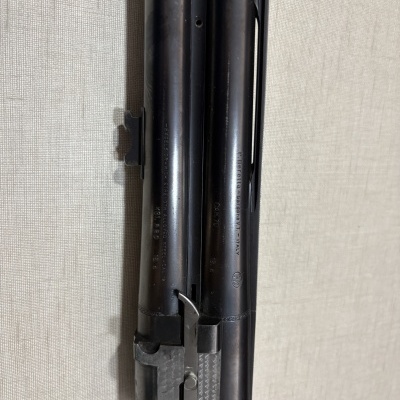 Cano duplo de arma de fogo negra com texto gravado em fundo tecido claro.
