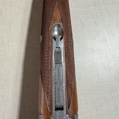 Arma de fogo com madeira castanha e metal gravado prateado sobre superfície bege.