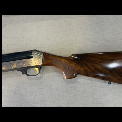 Espingarda de caça com coronha em madeira polida e detalhes em metal prateado com gravações douradas.