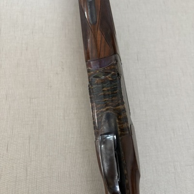 Arma de fogo com cabo em madeira escura e detalhe em metal marmoreado sobre fundo bege.