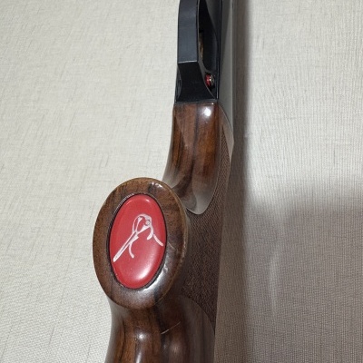 Espingarda com coronha em madeira castanha e detalhe oval vermelho com símbolo branco.