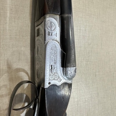 Espingarda sobreposta com metal gravado, madeira escura, gatilho duplo e fita vermelha sobre fundo bege