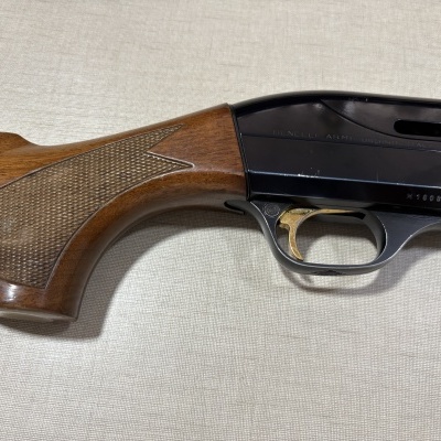 Parte da arma de fogo mostrando madeira, metal preto e detalhes em dourado com texto BENELLI ARMI URBINO ITALY
