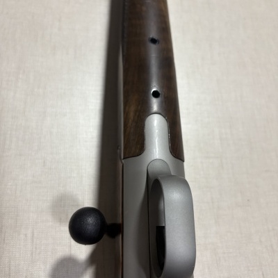 Parte traseira de espingarda ou rifle com madeira e metal cinza sobre fundo claro.