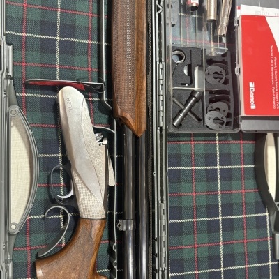 Estojo com espingarda Benelli, acessórios metálicos e manual vermelho sobre tecido xadrez.