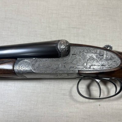 Espingarda com coronha em madeira escura e acabamento metálico gravado, com dois gatilhos e canos duplos.